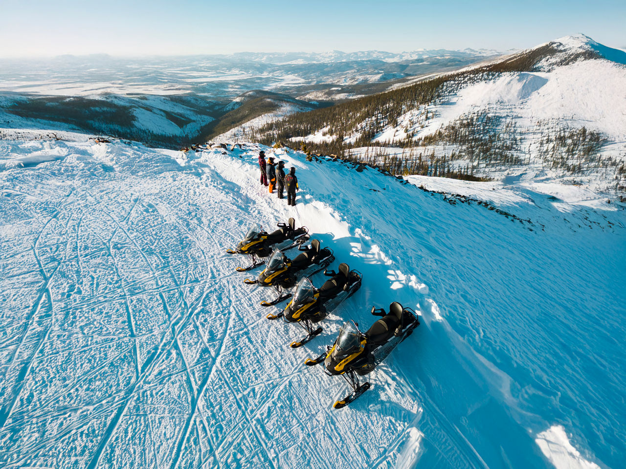 Vue aérienne de quatre personnes et quatre motoneiges Ski-Doo stationnées sur une crête de montagne enneigée sous un ciel bleu.