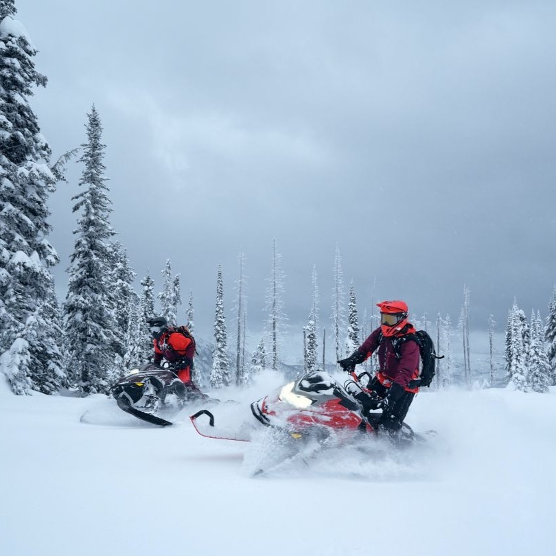 Two people riding BRP Lynx snowmobiles through a snowy mountain landscape.