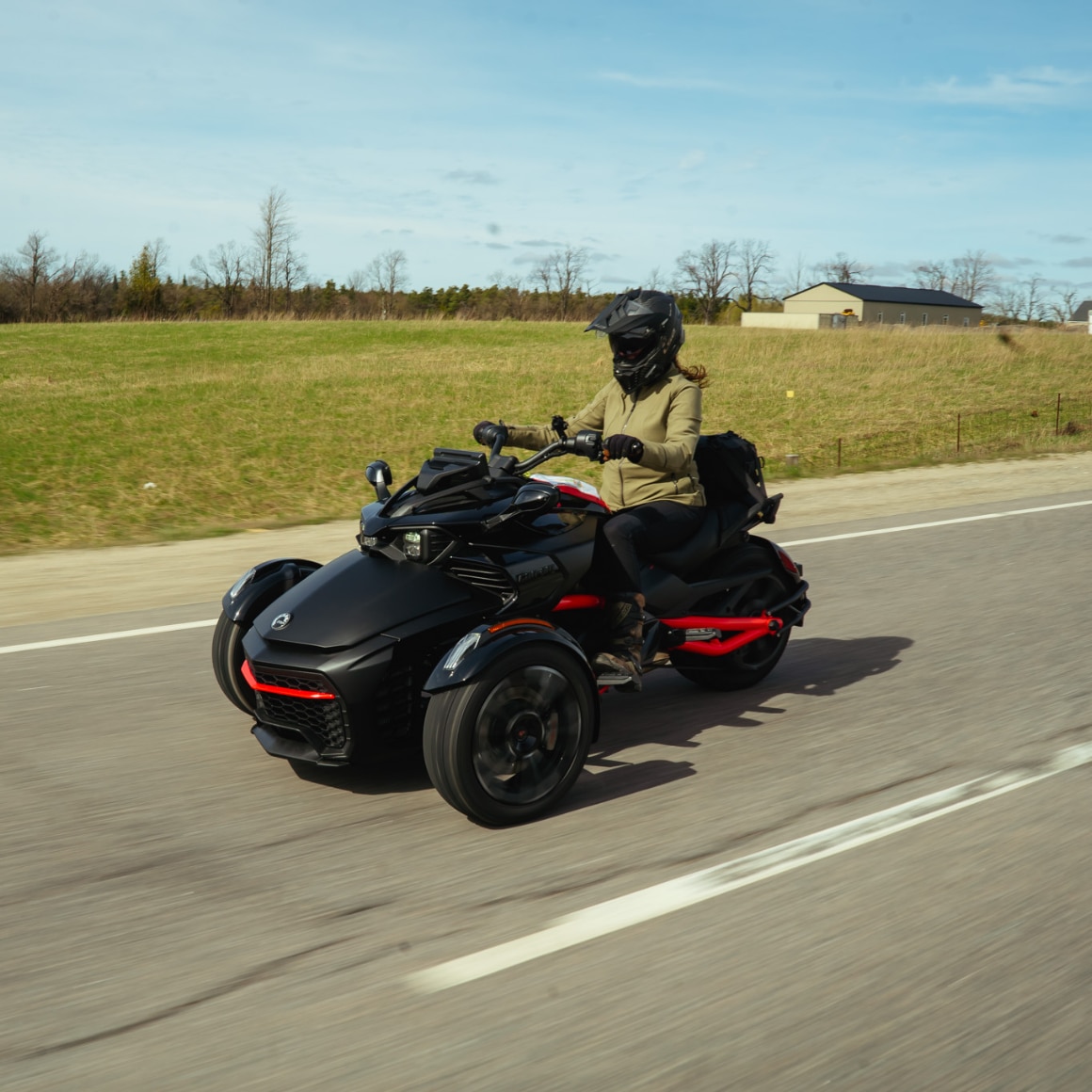 Woman riding a BRP Can-Am three-wheel vehicle on a sunlit paved country road.