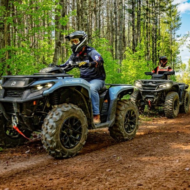 Two riders on BRP Can-Am ATVs traveling one behind the other through a sunlit forest trail during an adventure.
