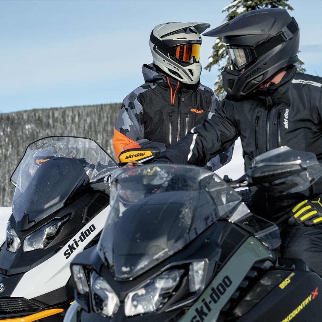 Two BRP Ski-Doo snowmobile riders in full gear conversing against a snowy mountain backdrop.